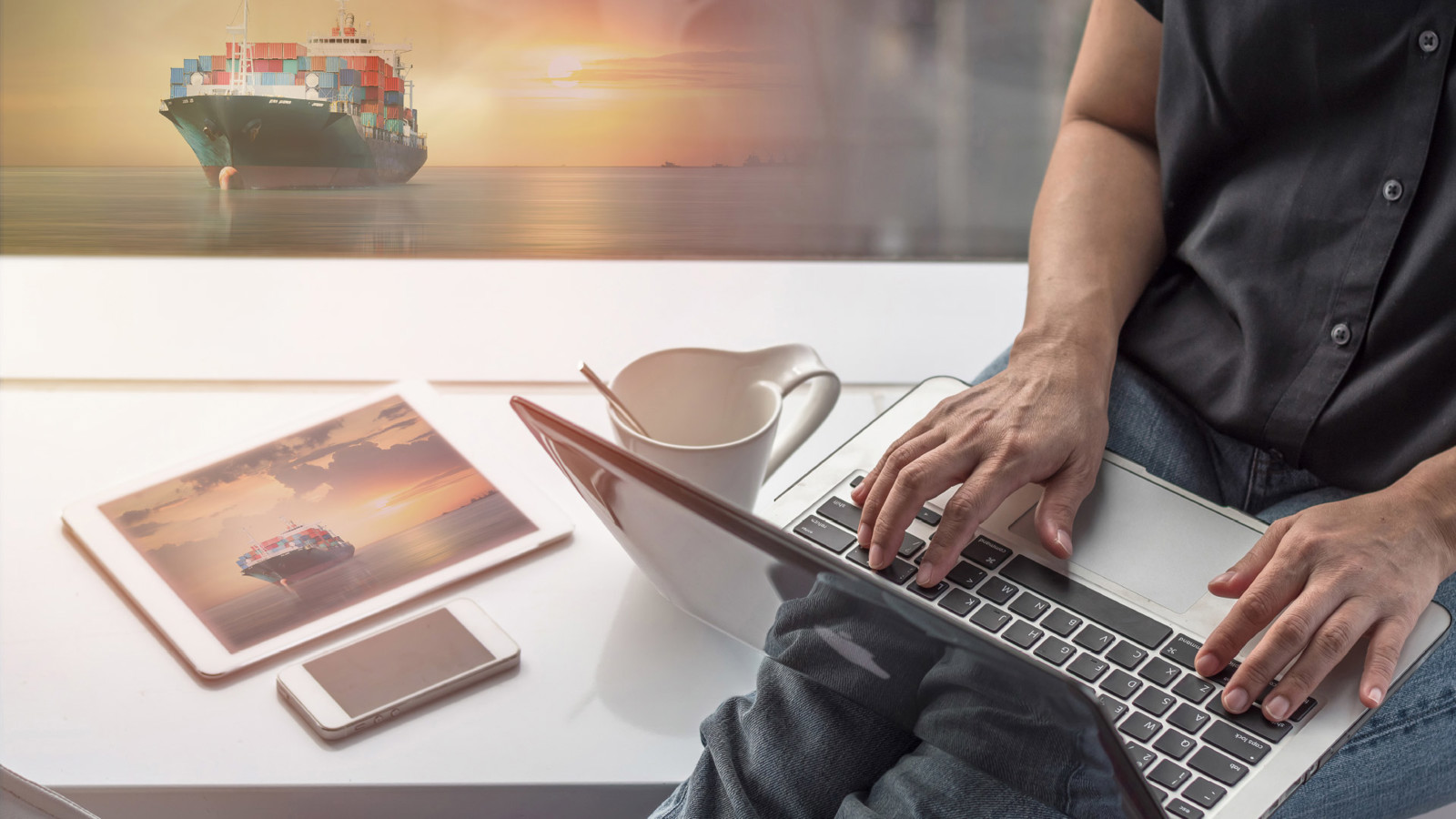 close-up of man sitting on a bench working on a laptop computer, tablet on the bench and the background shows a container ship on the sea with a sunrise/sunset on the horizon