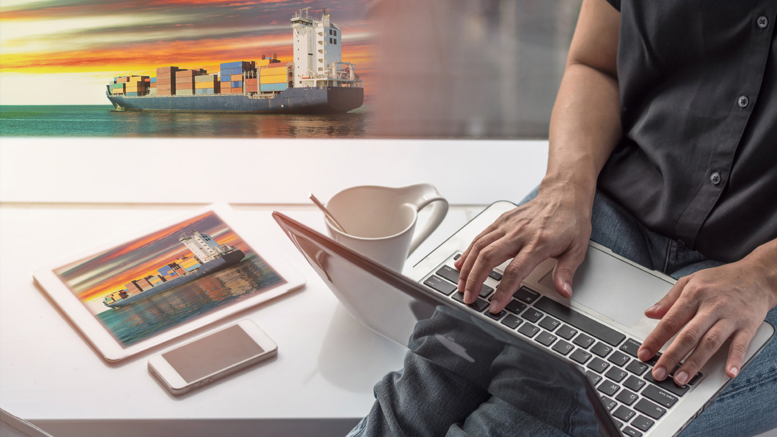 close-up of man sitting on a bench working on a laptop computer, tablet on the bench and the background shows a container ship on the sea with a vibrant sunrise/sunset on the horizon