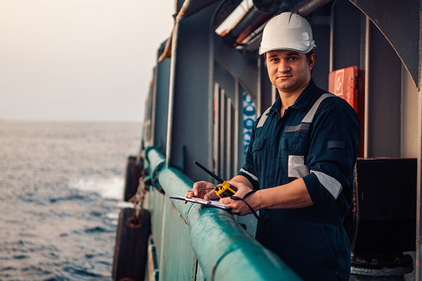 Deck officer standing at a ship's railing with a clipboard and walkie talkie.