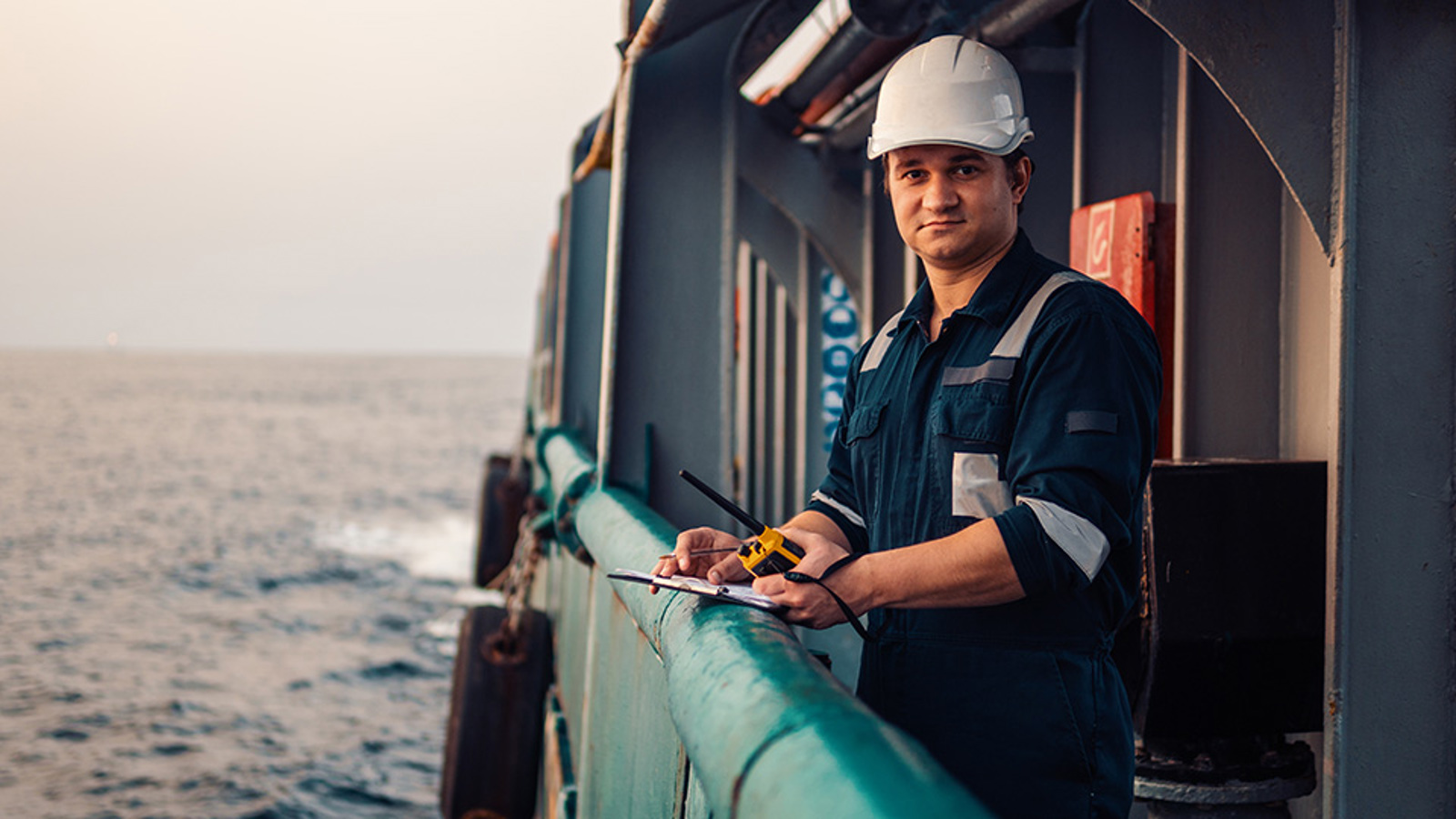 Deck officer standing at a ship's railing with a clipboard and walkie talkie.