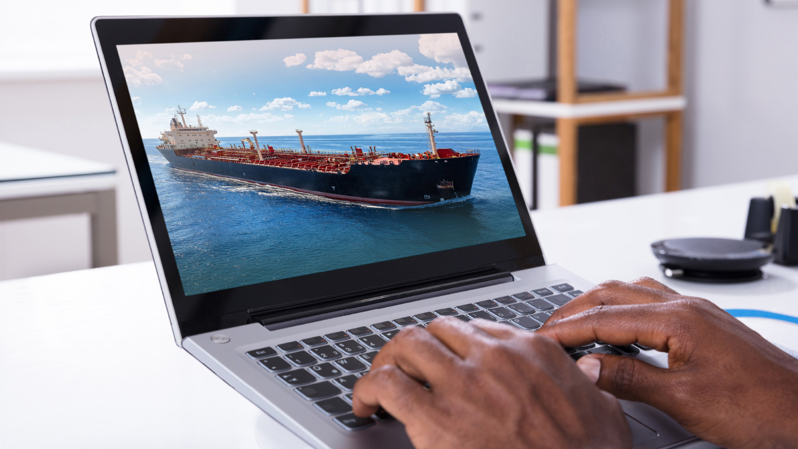 person's hands typing on a laptop computer showing a red & black tanker ship at sea on the screen